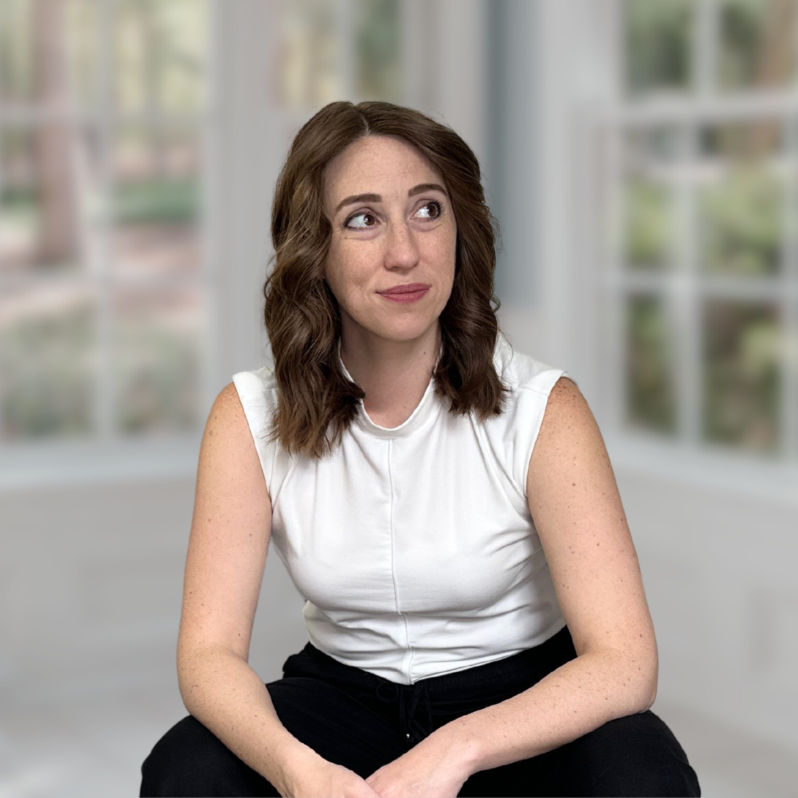 Image of Megan in a white top and black pants sitting in a room with windows in the background.