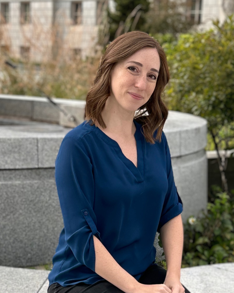 Photo of Megan in a blue top and black pants sitting outside on a concrete barrier.